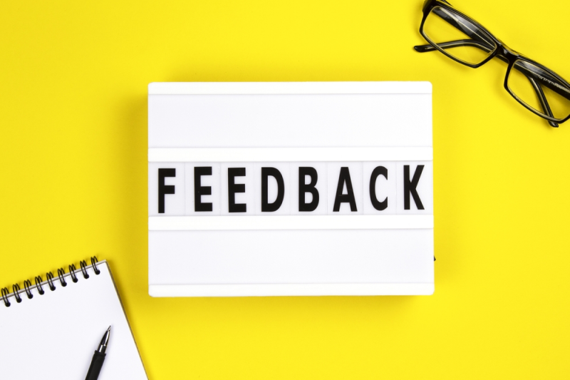 Letreiro branco com a palavra “Feedback” em letras pretas sobre fundo amarelo, ao lado de um caderno e óculos — representando o conceito de comunicação e retorno construtivo.