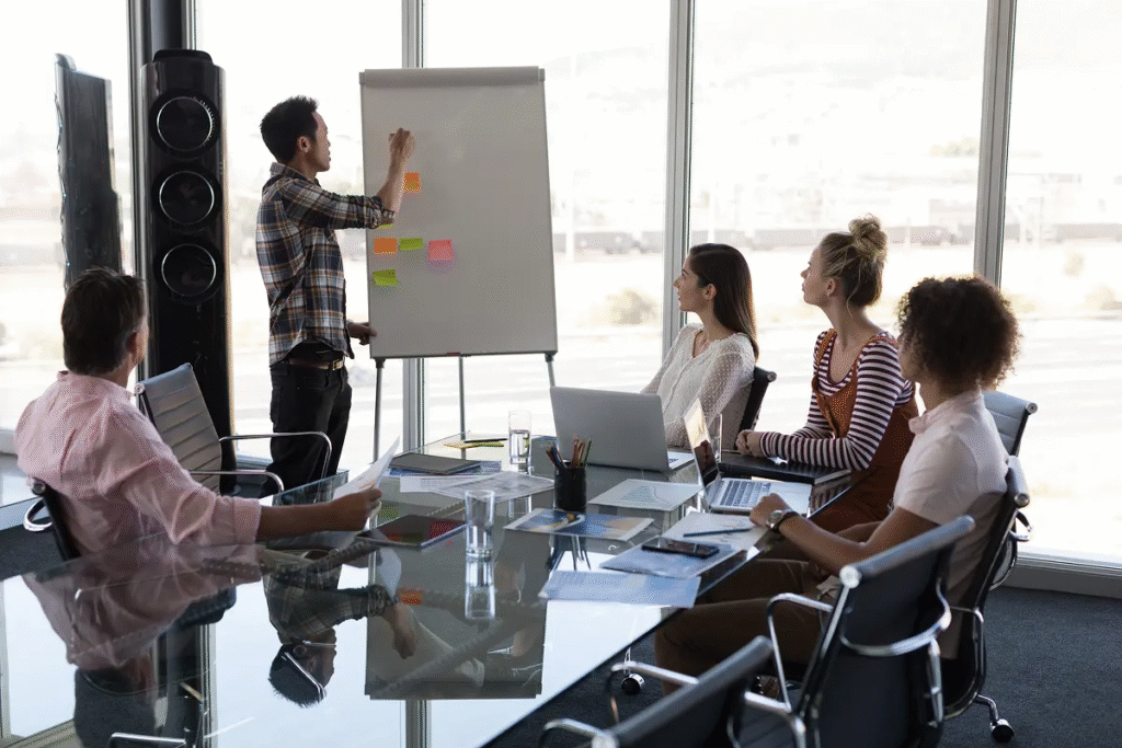 Grupo de profissionais em uma reunião de trabalho, com um líder apresentando ideias em um quadro branco enquanto os colegas observam, simbolizando o excesso de encontros corporativos e a importância de otimizar o tempo para aumentar a produtividade.
