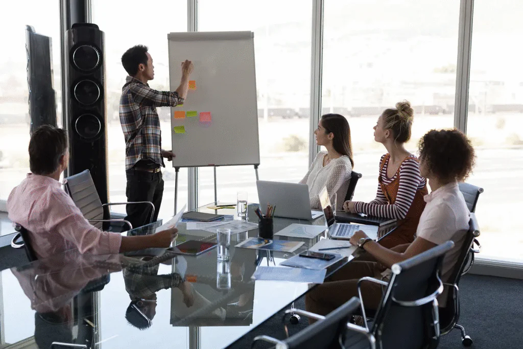 Grupo de profissionais em uma reunião de trabalho, com um líder apresentando ideias em um quadro branco enquanto os colegas observam, simbolizando o excesso de encontros corporativos e a importância de otimizar o tempo para aumentar a produtividade.